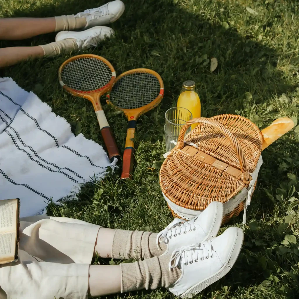 Two people lounging on grass in white sneakers after tennis, with a picnic basket, drinks, and a book creating a peaceful summer scene.