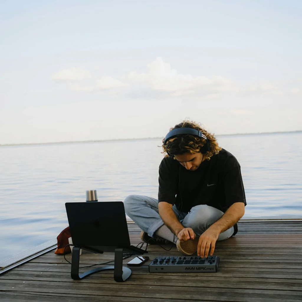 A person sits cross-legged on a wooden dock overlooking a calm body of water, working with music production equipment. The scene is captured during daylight with a light blue sky and scattered clouds and it shows active creative work in a peaceful setting.