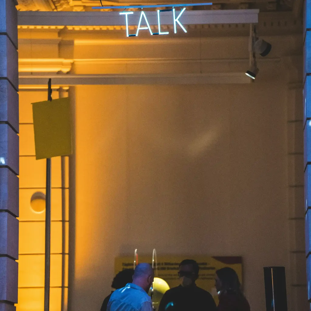 A neon 'TALK' sign illuminates people chatting on a balcony at night, champagne glasses visible in the warm amber lighting.