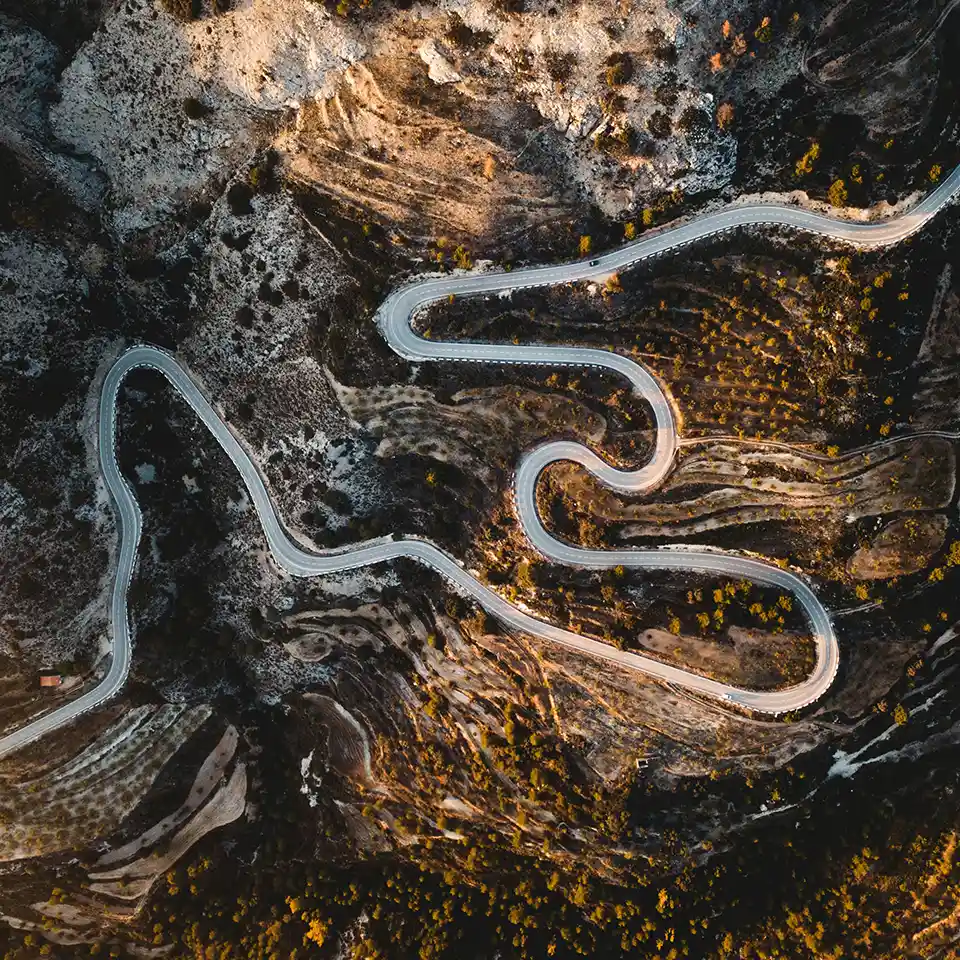 Drone shot of a narrow asphalt road with sharp turns winding through a dry, mountainous region with scrub vegetation.